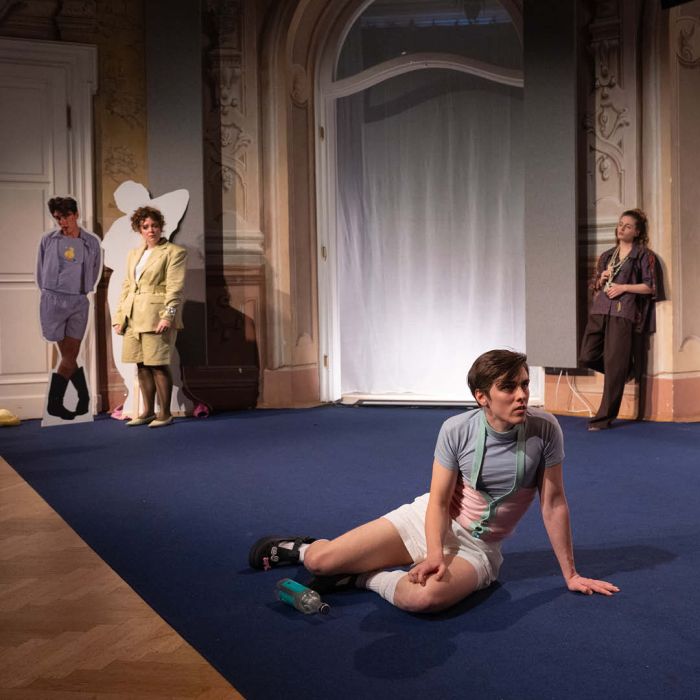 Am Boden sitzt ein Man seitlich, er trägt eine kurze weiße Hose. Dahinter stehen zwei Personen, die zu ihm blicken. IM Hintergrund, ein Türrrahmen mit einem weißen Vorhang.