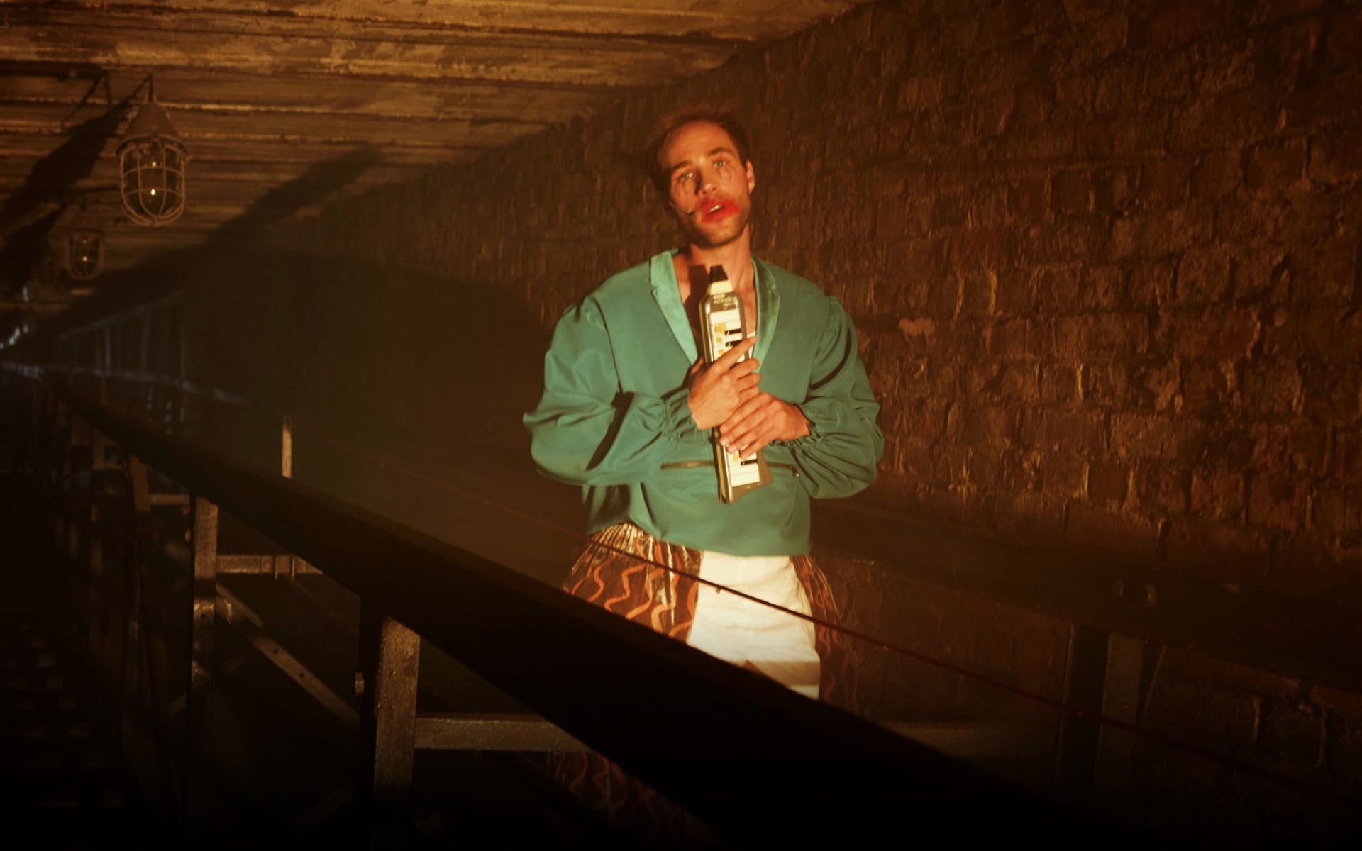 Ein Mann mit einer grünen Jacke hält eine Mundharmonika, sein Blick ist traurig. Er steht in einem dunklen Fabrikgelände und seine Lippen sind rot geschwinkt.