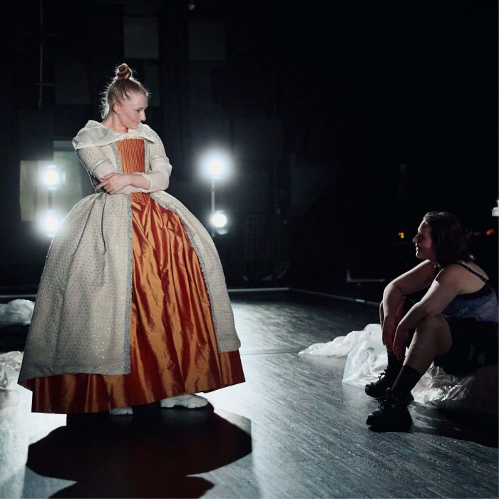 Zwei PErsonen blicken einander an, eine sitzt, die andere steht. Sie trägt ein historisches Kostüm.