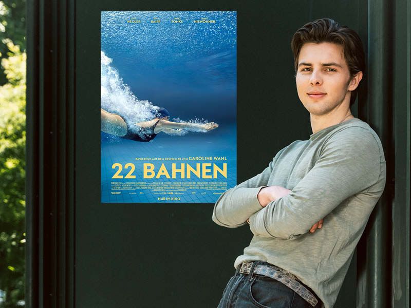 Ein junger Mann in einem hellgrünen langärmeligen T-Shirt lehnt an einer Wand, dahinter ein Plakat des Films "22 Bahnen"