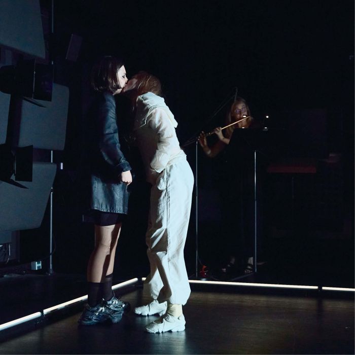 Zwei Personen küssen sich ungelenkt, die eine ist schwarz gekleidet, die andere weiß - im Hintergrund spielt eine Frau Querflöte-