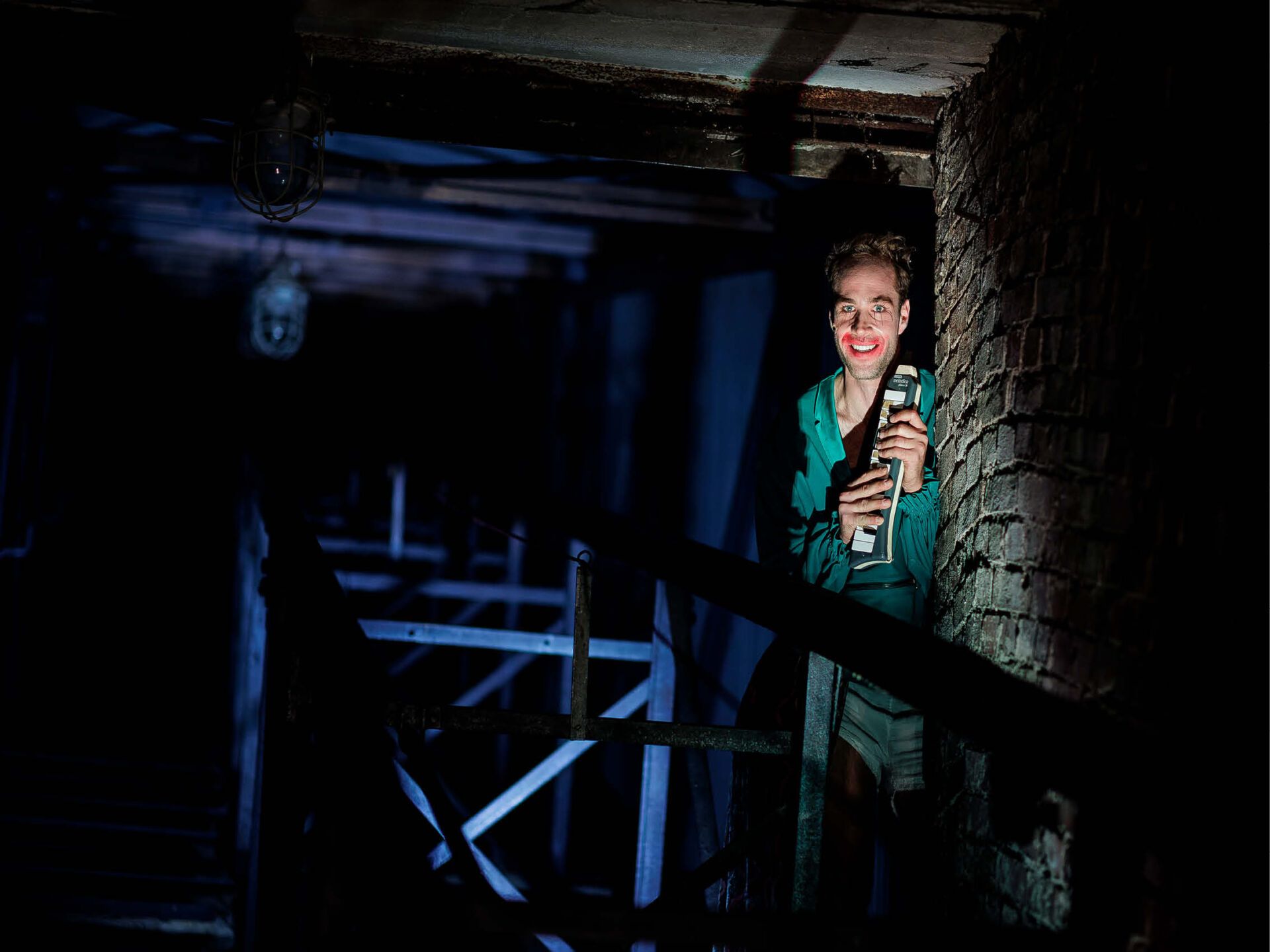 In einem dunklen Fabriksgelände mit Ziegelwand steht ein Mann mit grüner Jacke. Seine Lippen sind stark überschminkt. er grinst breit und hält eine Mundharmoniak in Händen.