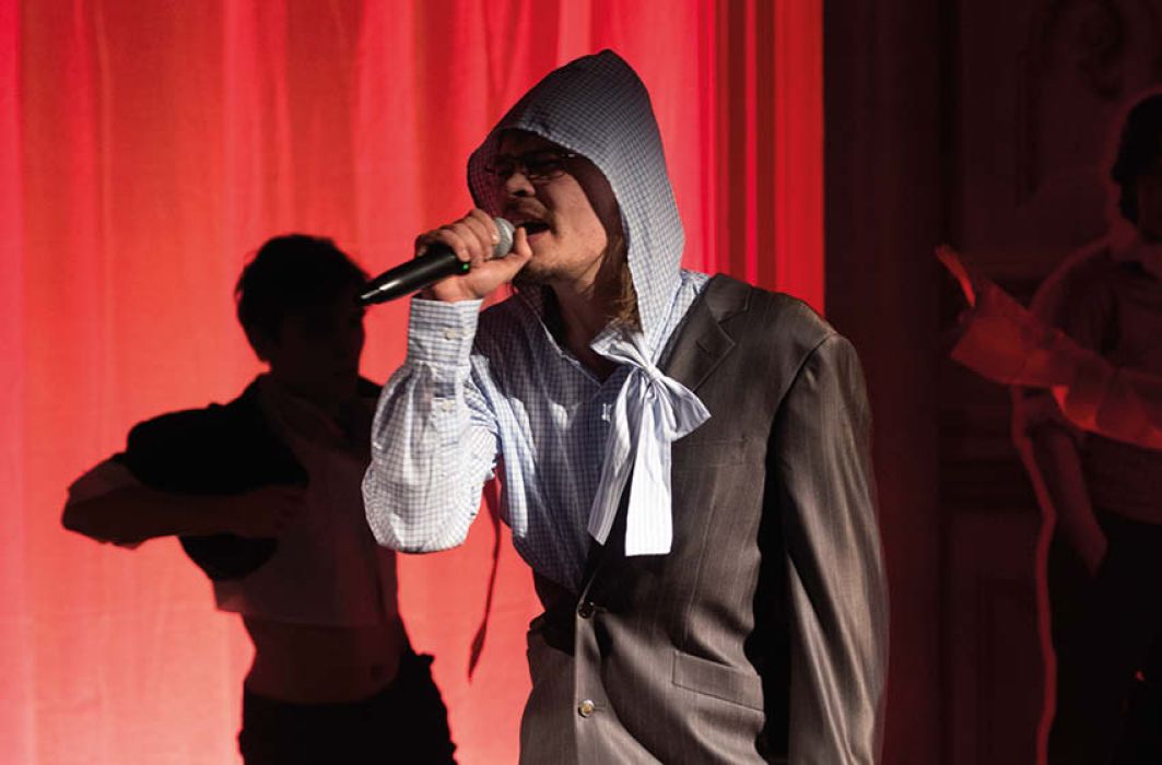 Eine Person mit grauem Sakko, das über die eine Schulter hängt und Kapuzenhemd sind, während hinten jemand tanz. Sie Szenerie ist in rotes Licht getaucht.