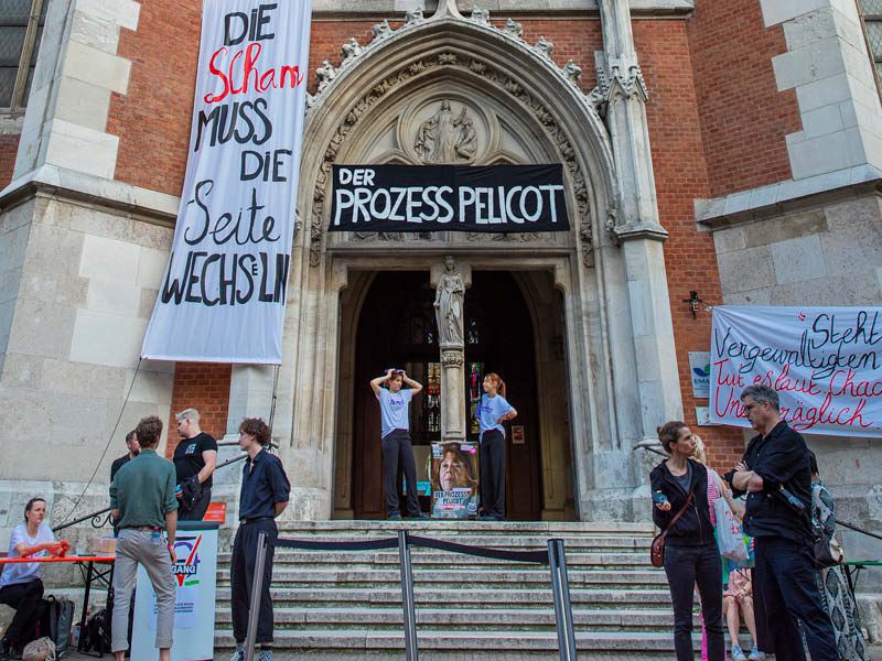 Ein Kirchenportal vor dem ein paar Menschen stehen, darauf Transparente mit "Der Prozess Pellicot" und "Die Scham muss die Seite wechslen"