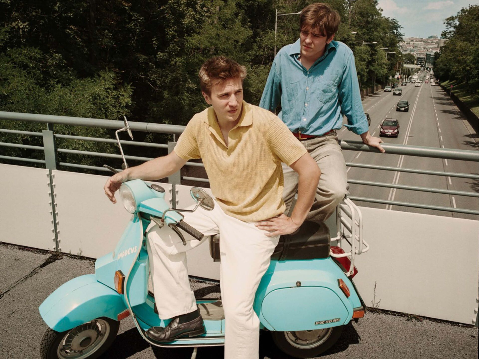 Zwie junge Männer stehen auf einer Brücke über eine mehrspurige Straße. Einer lehnt am Geländer, der andere sitzt auf einer türkisblauen alten Vespa.