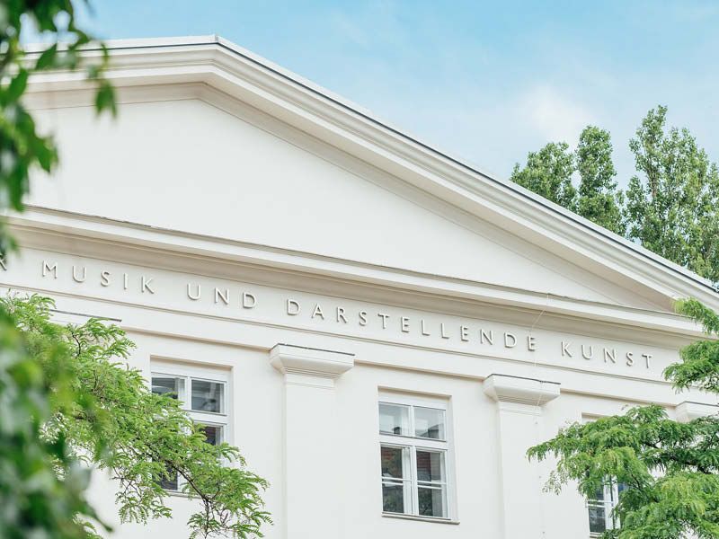 Zwischen ein paar Ästen mit grünen Blättern sieht man den First des historischen Teils der mdw, darauf ist mit goldenen Lettern der Name der Universität angebracht.