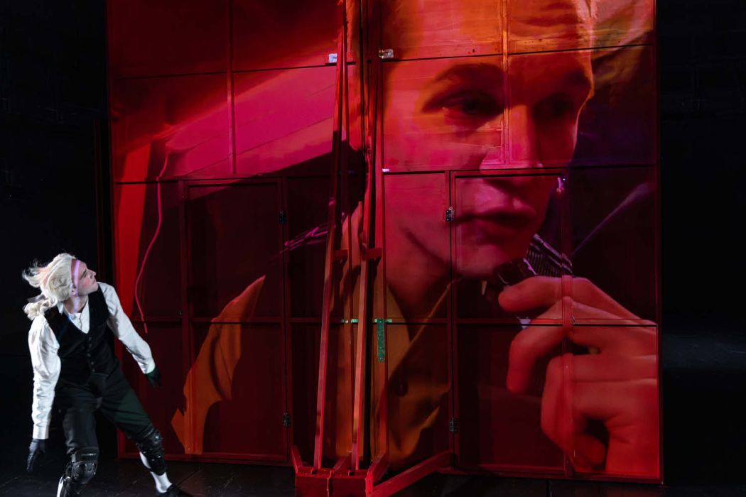 Auf der Videoeinspielung sieht man in rot eine Projektion einer Figur, links unten blickt eine Figur in historischem Kostüm nach oben - Perücke.