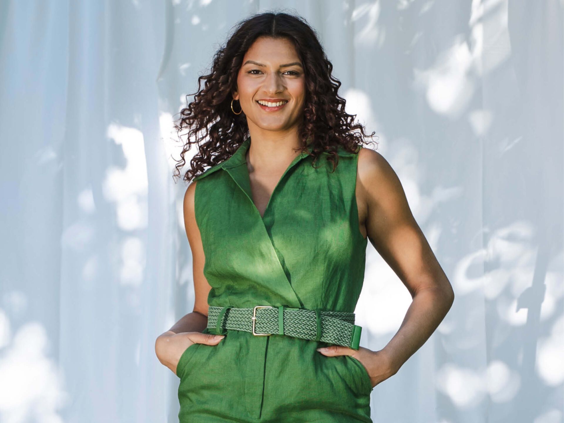Eine Frau mit dunklen Locken blickt lächelnd in die Kamera. Sie trägt einen grünen Jumpsuit und hat die Hände in die Hosentaschen gesteckt.