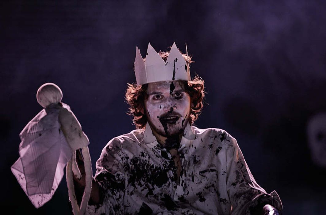 Eine Person hält eine kleine weiße gesichtslose Stoffpuppe in der Hand, die Person grinst, trägt eine Krone, das Gesicht ist weiß geschminkt, überall klebt schwarze Farbe.