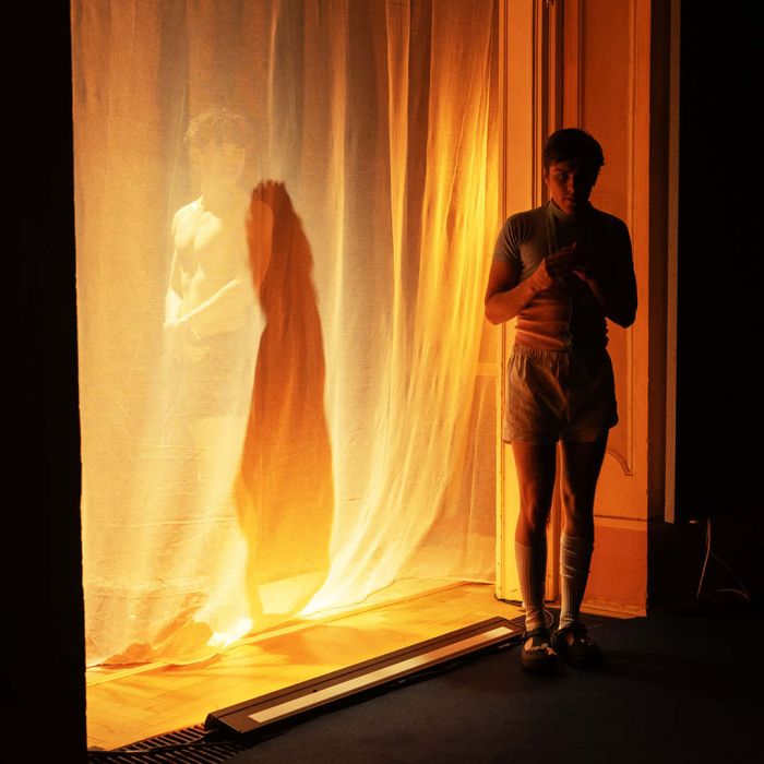 Hinter einem Semintransparenten Vorhang steht ein halbnackter Mann, der kaum zu sehen ist. Davor steht eine zweite PErson, die nach untern Blick. Das Licht taucht die Szenerie in gelbe Farbe.