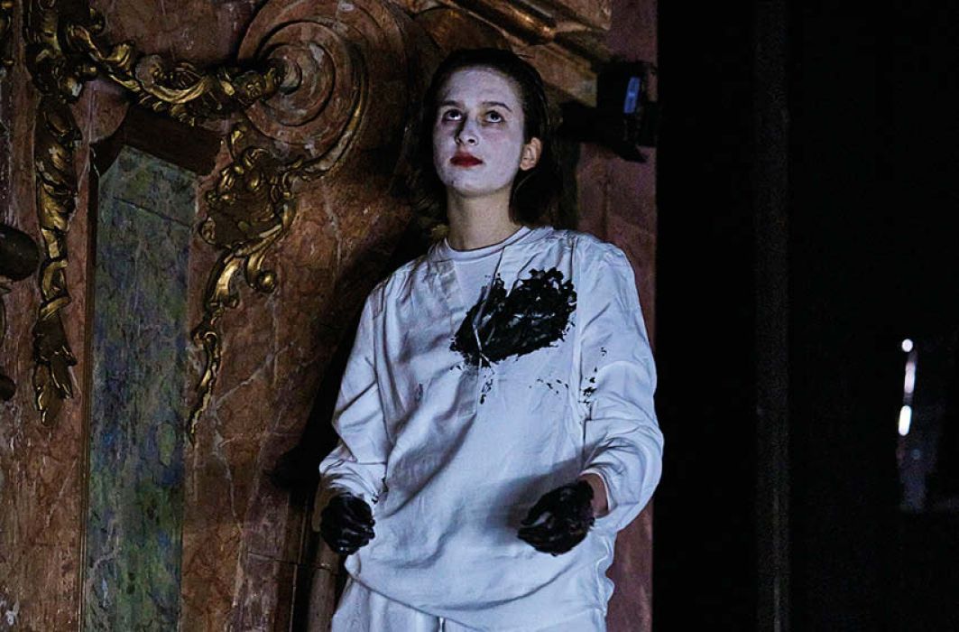 Eine Person lehnt an einem historischen Hintergrund, sie trägt weiße Kledung, das Gesicht ist weiß geschminkt, ihre Hände sind schwarz, überhalb ihrer linken Brust ist ein großer schwarzer Fleck.