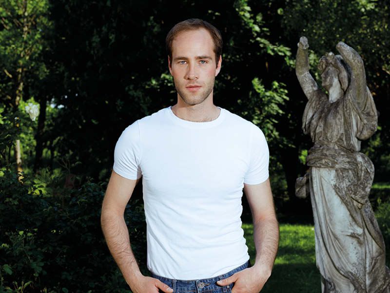 Fotos von Flo Sohn im Garten - er trägt ein weißes T-Shirt und Jeans, Blick in die Kamera, die Statue neben ihm hat keine Händer.