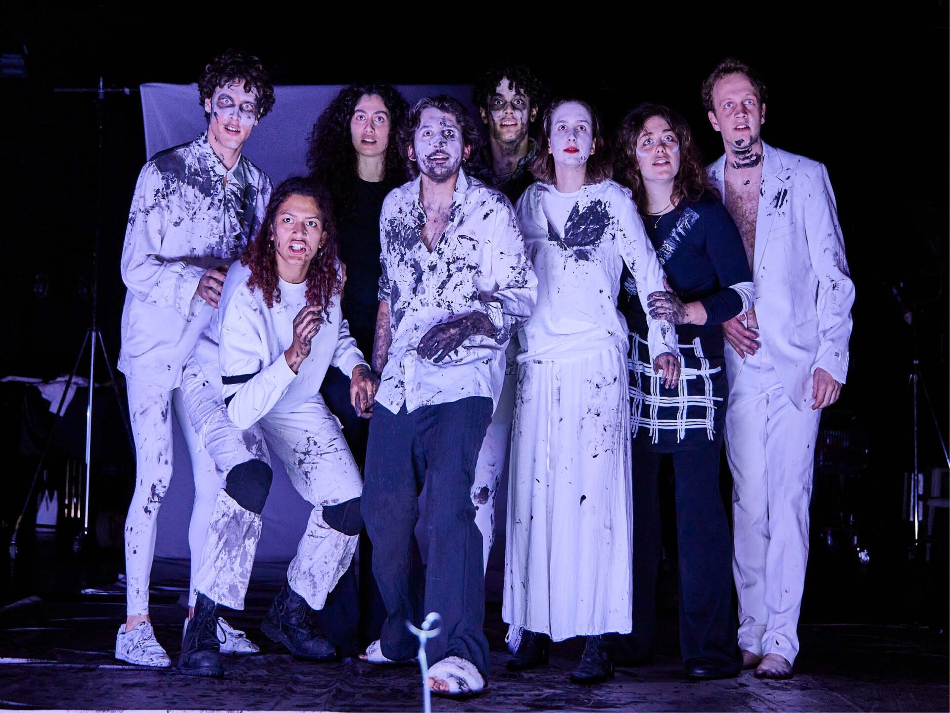 Die Gruppe spricht im Choral und blickt erstaunt, alle sind in weiß mit einigen schwarzen Akzenten gekleidet: Sie haben schwarze Farbe an Körper, Händen und im Gesicht.