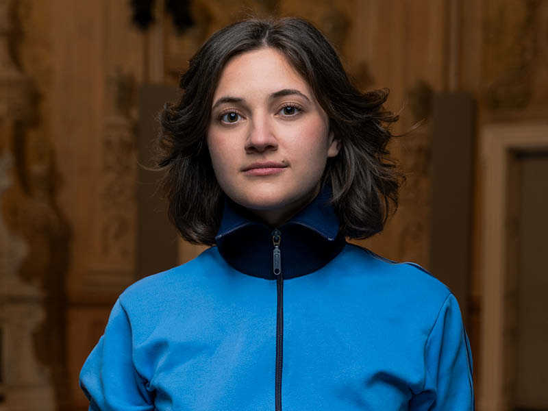 Eine Frau steht vor einem dunklen Hintergrund, sie hat braune Haare und lächelt in die Kamera. Sie trägt eine blitzblaue sportliche Jacke.