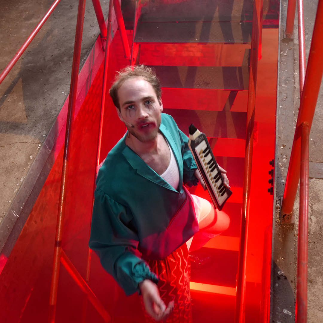 Auf einer Treppe steht ein Mann mit einer türkis-grünen Jacke, er hält eine Mundharmonika in der Hand.. von unten sind die Stufen in rotes Licht getaucht.