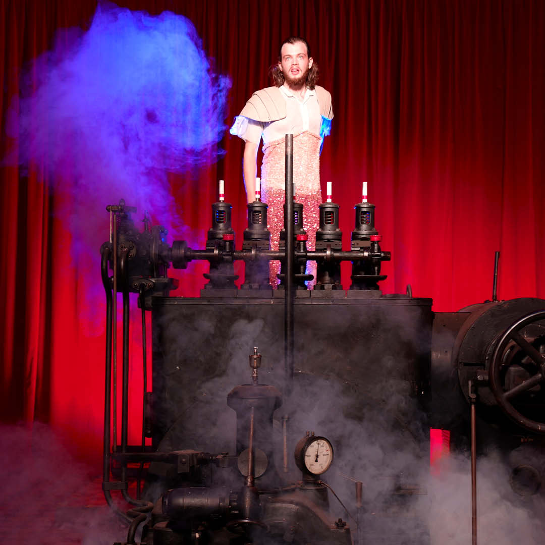 Ein Mann steht an einer großen dunklen Maschine mit mehreren Reglern, er ist weiß gekleidet. hinter ihm ein roter Vorhang und vor ihm etwas blauer Nebel.